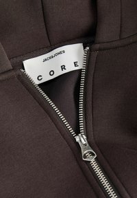 Fermeture éclair argentée partiellement ouverte sur une veste marron foncé montrant une étiquette en tissu blanche avec le nom de la marque Jack & Jones CORE.