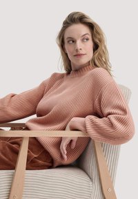 Strickpullover in sanftem Koralle mit Rippstruktur, hohem Kragen und locker sitzenden Ärmeln. Lässig getragen, während er in einem hellen Stuhl sitzt.