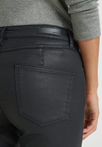 Svarta denimjeans med en slät yta, en bakficka med triangulär form och en subtil läderpatch på midjebandet.