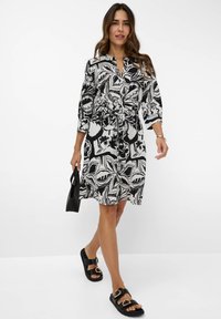 Schwarz-weiß gemustertes Kleid mit Dreiviertelärmeln, tailliertem Schnitt und knielanger Länge, kombiniert mit schwarzen Sandalen und einer kleinen schwarzen Tasche.