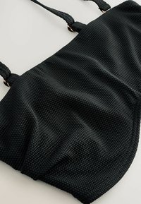 Čierna texturovaná látková taška cez rameno s pletenými popruhmi pripevnenými zlatými kovovými krúžkami, zobrazená na svetlom pozadí.