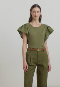 Top verde oliva con maniche a sbuffo, abbinato a pantaloni verdi oliva su misura e una cintura marrone con una fibbia circolare metallica.