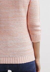 Pull en maille rose clair à manches trois-quarts porté avec un jean bleu foncé, vu de dos de la taille jusqu'aux hanches.