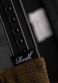 Brown textured fabric with vertical stripes, featuring a black tag with white cursive "Reell" text, attached to a black object.