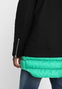 Osoba ubrana w czarną kurtkę ze srebrnym zamkiem na rękawie, na jasnej zielonej warstwie puchowej i niebieskich dżinsach, widoczna od tyłu.