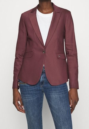 Persoon in een nauwsluitende bordeauxrode blazer over een wit shirt en blauwe jeans, staand tegen een effen achtergrond.