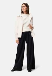 Ivory nep bontjas met oversized kraag, knoopaccenten, crèmekleurige geribde trui en brede zwarte broek, compleet met zwarte schoenen.