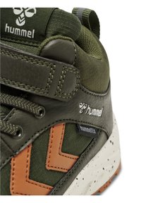 Baskets vertes avec des accents marron, présentant un dessus en cuir texturé et en tissu, une sangle réglable et une semelle blanche marbrée. Logo affiché sur le côté.