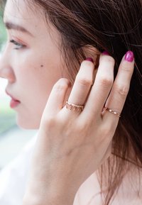 Anillos de oro rosa llevados en los dedos, uno con acentos colgantes y otro con un diseño entrelazado. Textura suave, brillo sutil.