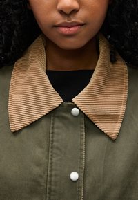 Εγγύς λήψη ενός ατόμου που φοράει παλτό ελιάς με μπεζ ριγωτό γιακά από κορντόρυ και ασημένια κουμπώματα με κλικ, εμφανίζοντας μόνο το κάτω μέρος του προσώπου και το λαιμό.