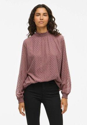 Femme aux cheveux bruns ondulés portant un chemisier rose texturé à manches longues et un pantalon noir, debout devant un fond clair uni.
