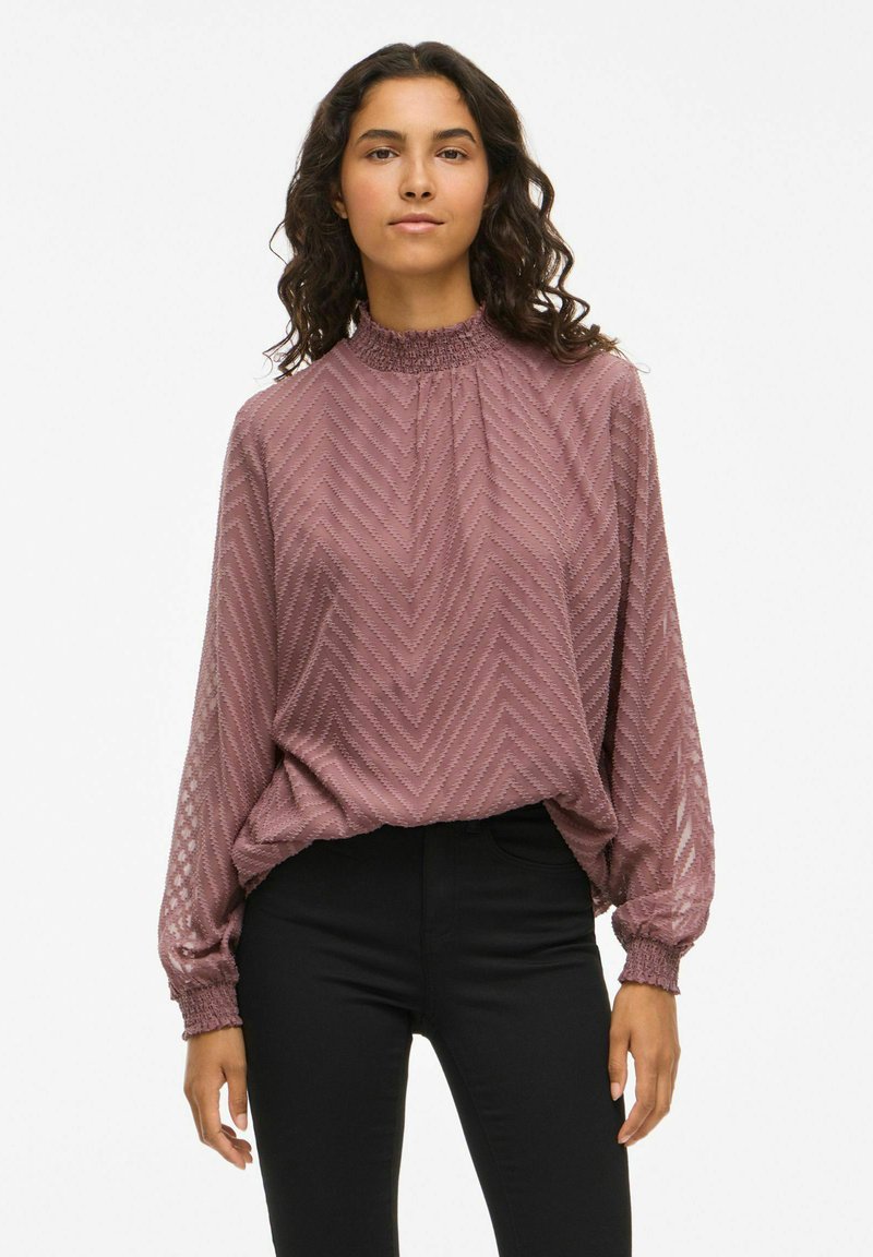 Femme aux cheveux bruns ondulés portant un chemisier rose texturé à manches longues et un pantalon noir, debout devant un fond clair uni.