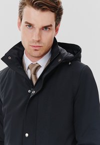 Czarna wodoodporna kurtka z kapturem, zamek błyskawiczny oraz zatrzaskowe guziki; charakteryzuje się gładką teksturą i prostymi liniami, w połączeniu z wzorzystym krawatem.