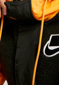 Svart och orange jacka med fleece-textur, med en orange dragsko, en svart zippered sektion och en vit Nike-logotypdetalj.
