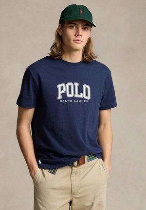 Joven con gorra verde oscuro, camiseta azul marino de "Polo Ralph Lauren", pantalones beige y cinturón a rayas, de pie con las manos en los bolsillos.