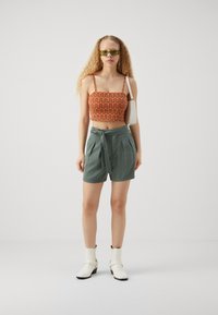 Kratká oranžová blúzka s vlnitým vzorom; zelené šortky so šnúrkou; biele členkové čižmy. Modelka nosí zelené slnečné okuliare a má kučeravé vlasy.