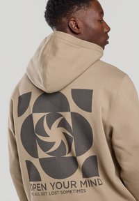 Beige hoodie with a large black graphic print on the back featuring abstract shapes and text reading "OPEN YOUR MIND." Cotton material, relaxed fit.