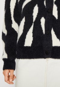 Pletený cardigan s černo-bílým abstraktním vzorem, vyrobený z chlupatého materiálu, se zapínáním na knoflíky vpředu a žebrovanými manžetami a lemem.