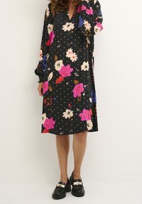 Svart blommig klänning med stora rosa, vita och blå blommor, med V-ringning och långa puffärmar. Matchas med kraftiga svarta skor.