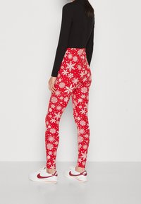 Rode leggings met een wit sneeuwvlokpatroon. Gemaakt van rekbare stof, strakke pasvorm. Gedragen met een zwart topje en witte sneakers met rode accenten.