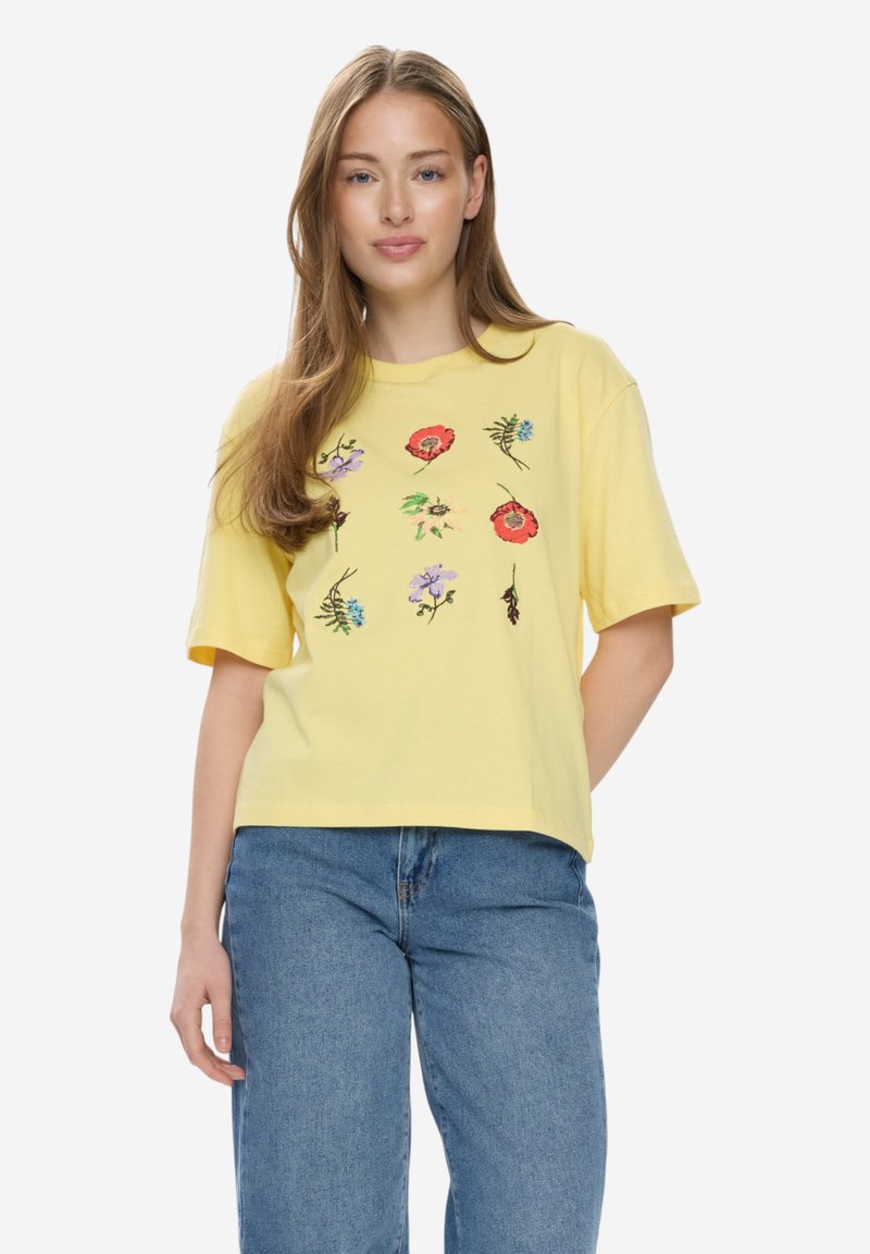 Jeune femme aux longs cheveux châtain clair portant un t-shirt jaune avec des motifs floraux colorés et un jean bleu, debout devant un fond blanc.