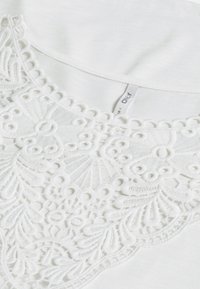 Vit T-shirt med intrikata vita broderier vid halsringningen, med vågiga kanter och en mjuk, texturerad finish.