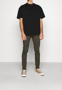 Czarna bawełniana koszulka o luźnym kroju, zestawiona z oliwkowozielonymi spodniami o dopasowanym kroju oraz brązowymi niskimi trampkami z białą podeszwą i sznurówkami.