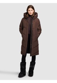 Brązowa puchowa kurtka z dużym kapturem, zamkiem błyskawicznym i pikowaną fakturą. Noszona na ciemnym swetrze, w połączeniu z czarnymi śniegowcami do połowy łydki.