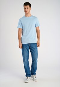 T-shirt in cotone azzurro chiaro con maniche corte, caratterizzata da dettagli di testo discreti, abbinata a jeans di denim blu e scarpe da ginnastica bianche.