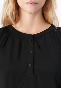 Lys blond kvinne iført en svart bluse med rund hals og tre svarte knapper nedover midten.
