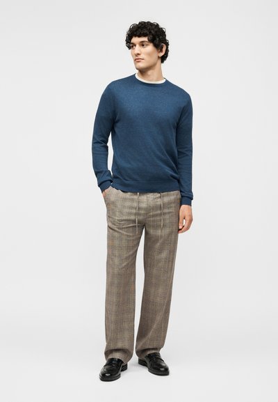 Ένας νεαρός άνδρας στέκεται με τα χέρια στις τσέπες, φοράει ένα μπλε πουλόβερ, μπεζ καρό παντελόνια και μαύρα δερμάτινα παπούτσια σε λευκό φόντο.