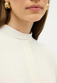 Crème blouse met een hoge hals, subtiel textuurpatroon, gecombineerd met grote, goudkleurige creolen. Close-up van de nek en het gezicht.