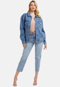 Lichtblauwe denimjacket met een ontspannen pasvorm, voorzien van grote zakken en knoopsluitingen, gecombineerd met lichtblauwe jeans en naakte bandjesheels.