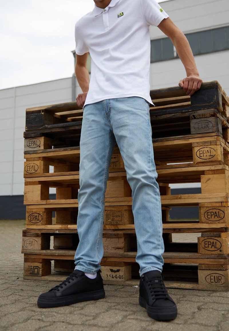 Weißes Poloshirt mit kleinem grünem Logo, hellblaue Jeans und schwarze Turnschuhe, stehend vor aufgestapelten Holzpaletten auf dem Gehweg.
