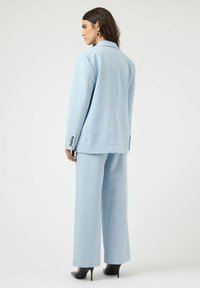 Γυναίκα με ανοιχτό γαλάζιο κοστούμι με παντελόνι φαρδύ και σακάκι, γυρισμένη προς τα πίσω, φοράει μαύρα ψηλοτάκουνα παπούτσια και μαύρα σκουλαρίκια, πάνω σε λευκό φόντο.