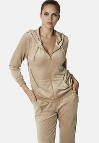 Ensemble de survêtement en velours beige comprenant un hoodie à zip avec cordon de serrage, des poches avant et un pantalon fuselé assorti avec un waistband élastique.