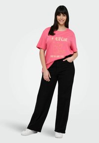 Różowa bawełniana koszulka z złotym napisem i grafiką, zestawiona z czarnymi szerokimi spodniami. Model nosi białe trampki. Płaskie tło.