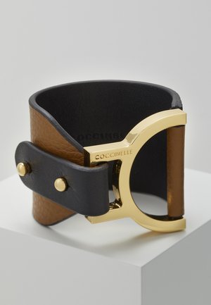 Szeroka skórzana bransoletka z brązowymi i czarnymi paskami, złotą sprzączką z wygrawerowanym napisem "COCCINELLE", zabezpieczona dwoma złotymi ćwiekami.