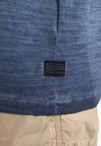 Námorně modrá látka s texturovaným povrchem, na dolním okraji se nachází černá tkaná etiketa "Petrol Ind". Jednoduchý, běžný design se střihy rovně.