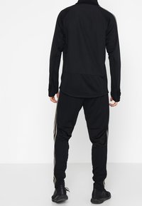 Schwarzer Sportanzug mit zip-up Oberteil und hohem Kragen sowie taillierten Hosen, akzentuiert durch seitliche Streifen in einer kontrastierenden Farbe.