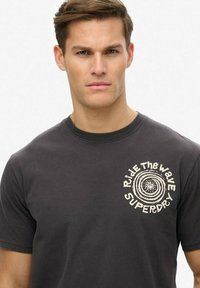 Camiseta de algodón gris con cuello redondo y mangas cortas, con un logotipo circular blanco y texto en el área del lado izquierdo del pecho.