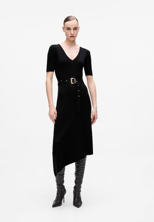 Abito nero lavorato a maglia con scollatura a V profonda, maniche corte e orlo asimmetrico. Presenta una vita con cintura e accessori metallici. Indossato con stivali neri al ginocchio.