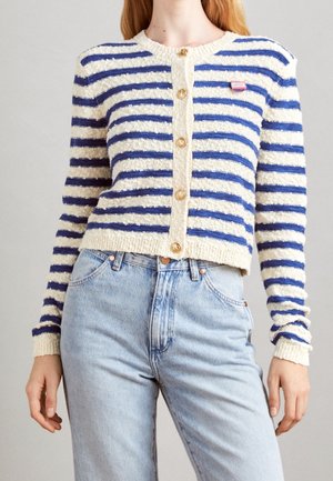 Femme portant un cardigan boutonné à rayures crème et bleues avec des boutons dorés et un jean taille haute bleu clair, debout devant un fond uni.