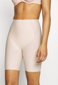 Beige high-waisted shapewear shorts gemaakt van soepel, rekbaar materiaal, met een naadloos ontwerp en een lengte tot halverwege de dijen, voorzien van een opening aan de voorkant.