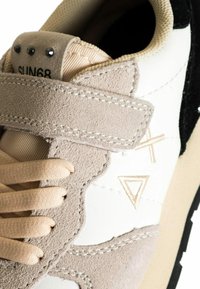 Prime piano di una sneaker beige e bianca con una cinghia in suede, lacci beige e dettagli di cucitura geometrica sul pannello laterale.