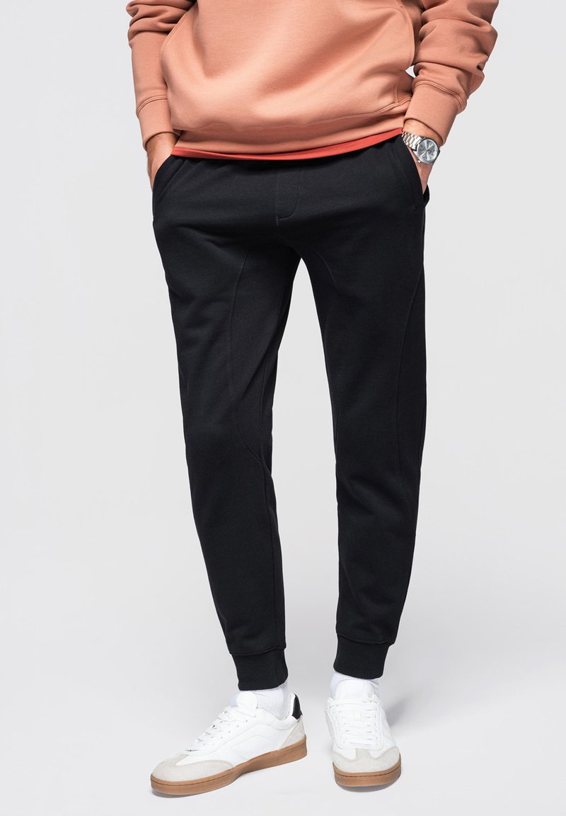 Persona vestida con pantalones jogger negros, zapatillas blancas y una sudadera color melocotón, con las manos en los bolsillos, de pie frente a un fondo neutral.