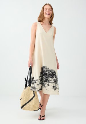 Femme portant une robe beige sans manches avec un imprimé village côtier noir, tenant un grand sac cabas tissé avec des anses noires, souriante et marchant.