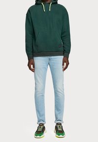 Sudadera verde con capucha y cordones amarillos, bolsillo frontal y dobladillo acanalado; combinada con jeans azul claro y zapatillas negro-verde.