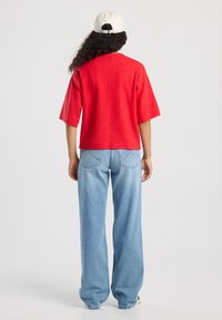 Henkilö, jolla on pitkät kiharat hiukset, valkoista lakkia, punainen lyhythihainen paita ja vaaleansiniset leveälahkeiset farkut, kääntyy pois yksinkertaista taustaa kohti.