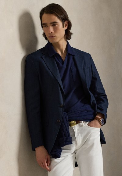 Νεαρός άνδρας ακουμπισμένος σε τοίχο, φορώντας ναυτικό μπλέ σακάκι, μπλε πλεκτό πόλο με πουλόβερ δεμένο στη μέση, λευκό παντελόνι και καφέ δερμάτινο ρολόι χειρός.